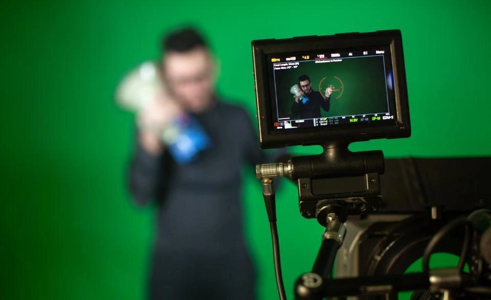 A video camera records a man, whose image is visible on the camera screen, while he stands in front of a green background used for green screen compositing (chromakey).